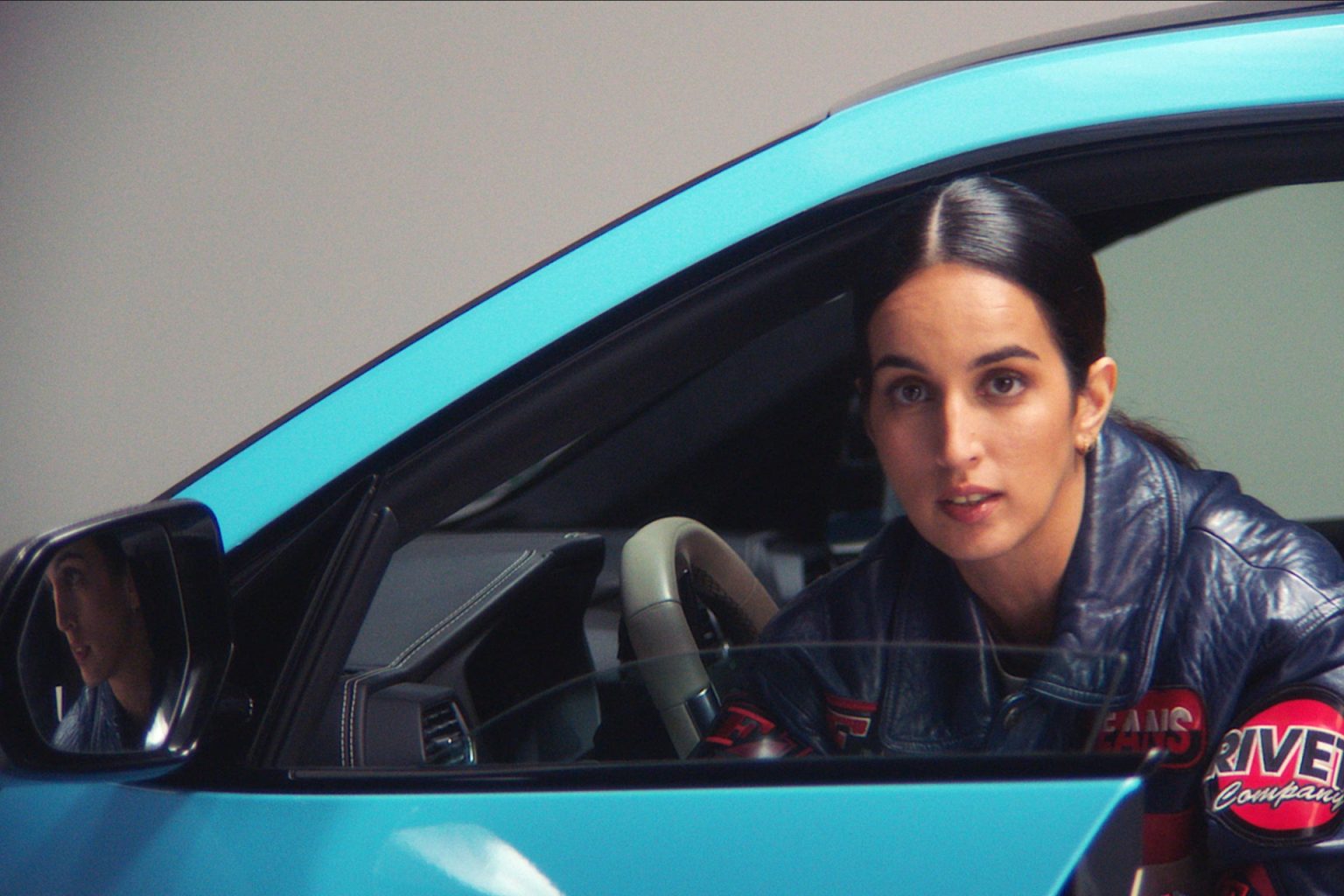 Auf dem Bild das geöffnete Fahrerinnensitzfenster eines hellblauen Autos zu sehen. Am Steuer sitzt die Rapperin Ebow und schaut aus dem Fenster in die Kamera. Sie trägt eine schwarze Lederjacke mit rotem Schriftzug und ihr glänzendes schwarzes Haar ist gescheitelt und zusammengebunden. Im Rückspiegel des Autos ist ihr Gesicht reflektiert.